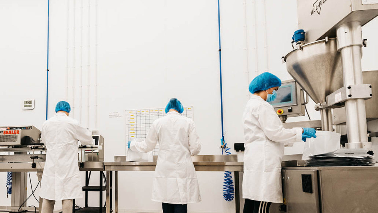 drie lijn medewerkers in witte overal en met haarkapjes en handschoenen, werken aan de vullijn in een supplementen productie.